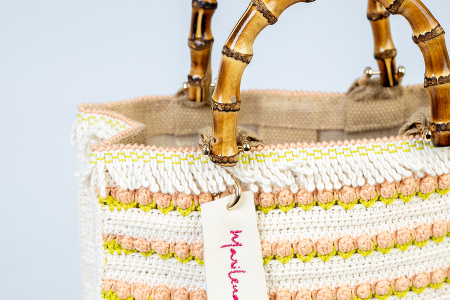 Incantevole Tote Bag composta interamente da ricercati dettagli, utilizzando il "Punto Tulipano". Piccoli fiori di un rosa pallido si alternano a linee di un bianco morbido. I manici in bambù, uno dei simboli identificativi del brand, permettono di indossarla come borsa a mano; La sua tracolla rimovibile, invece, permette di portare la borsa a spalla. Di manifattura artigianale, prodotta in Italia.